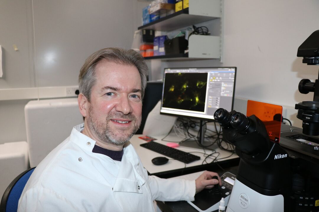 man wearing white lab coat sat next to large microscope 