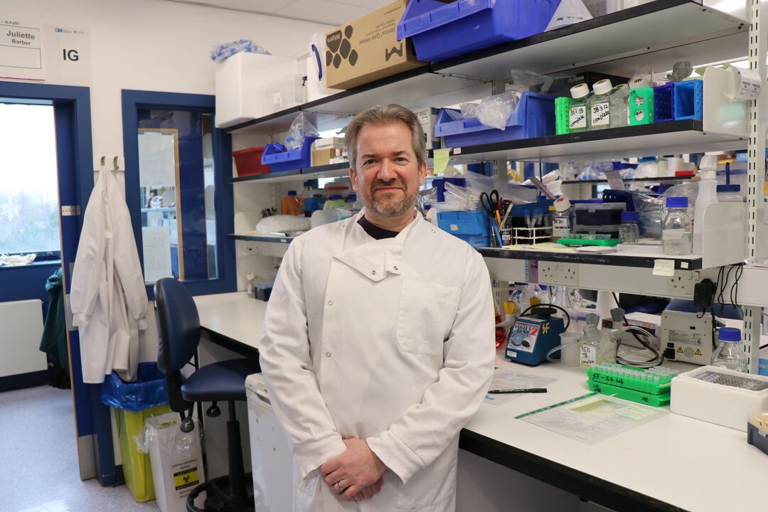 man wearing white lab coat sat standing in science lab leaning on bench with equipment behind him 