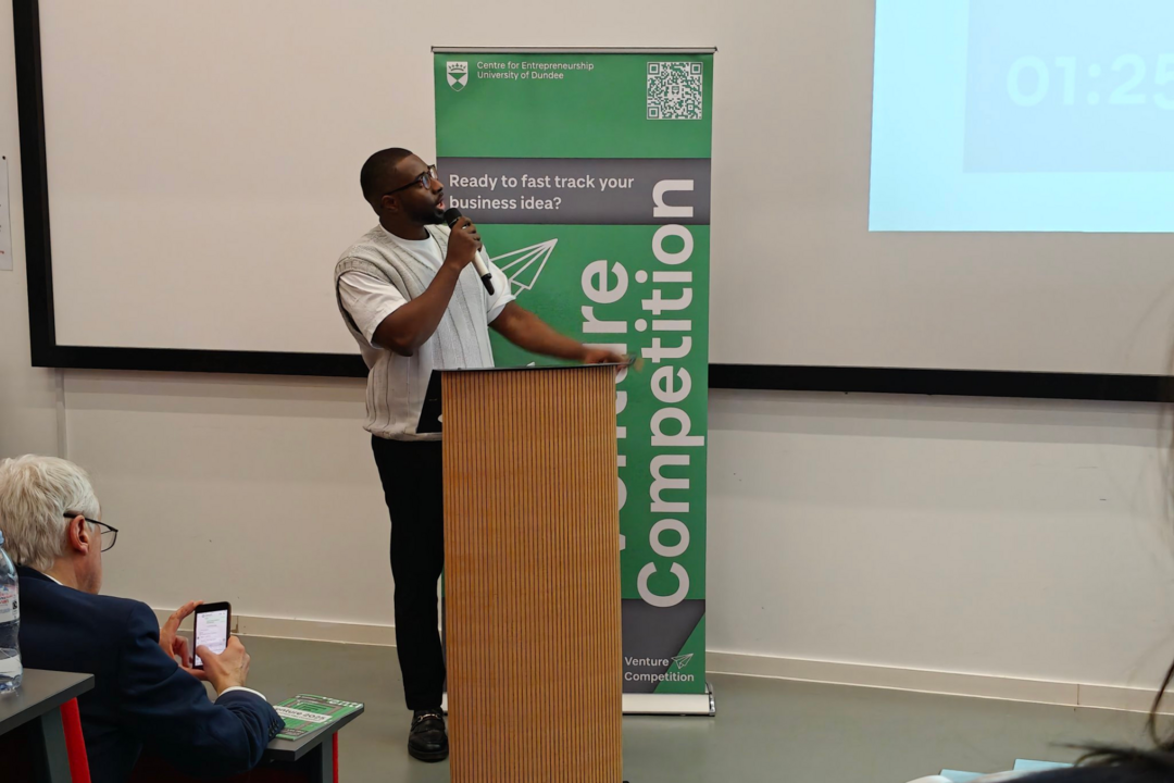 A man stands at a podium speaking into a microphone and looking up to the right at a projection on the wall.
