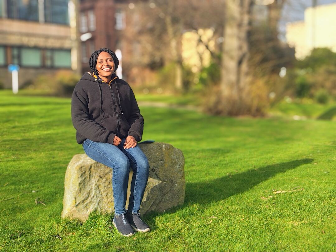 A woman sits on a boulder in a grassy space, smiling.