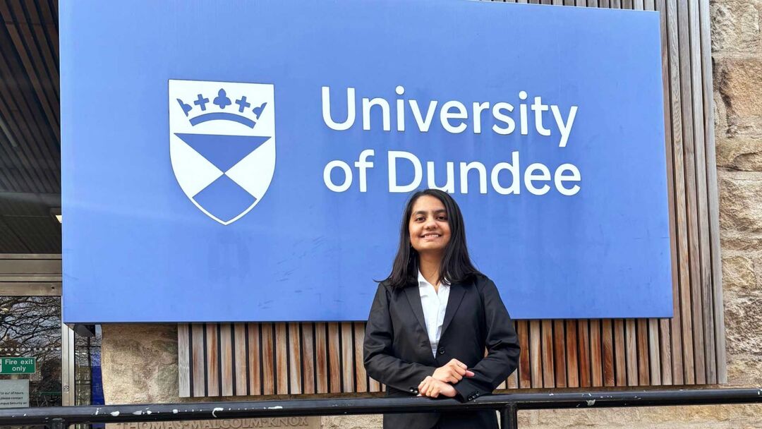 A person stands in front of a large blue University of Dundee sign with their hands resting on a metal rail in front.