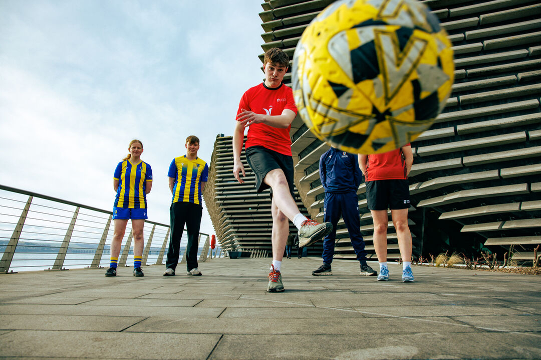 A group of people stand near the V&A building, one kicks a yellow football towards the camera.