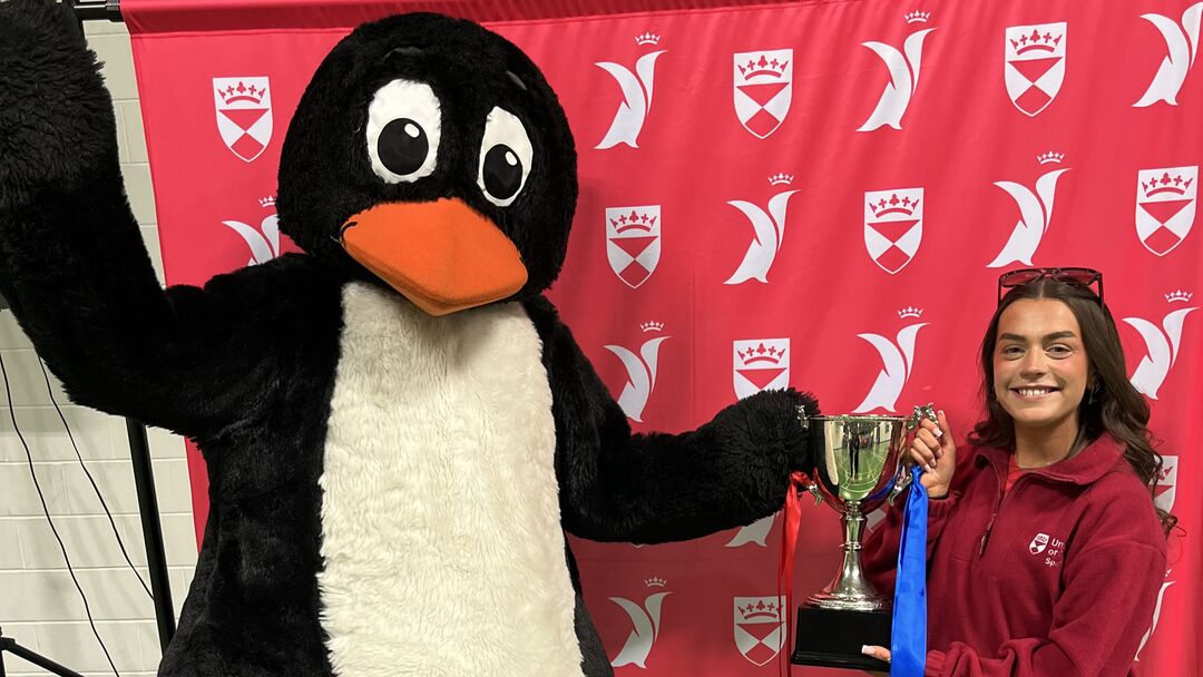 A person in a penguin mascot costume holds up a trophy with a girl with long brown hair.