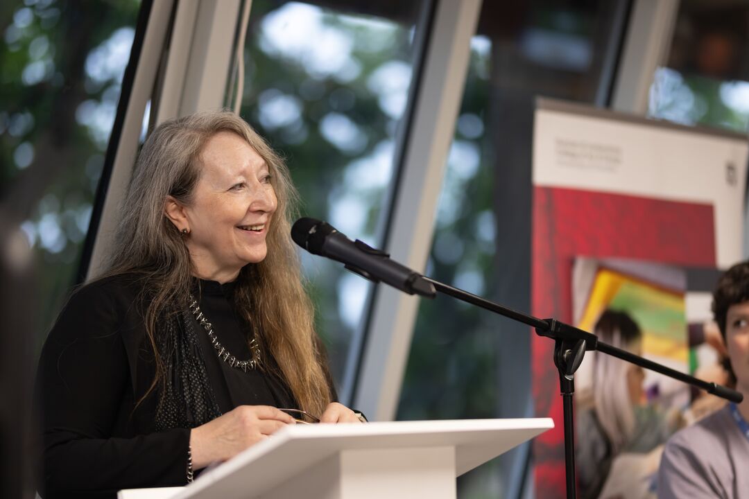 Woman with long hair standing behind a speaking podium, with a microphone in front of her, mid-speech and smiling
