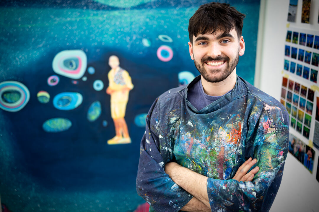 Young man with dark hair and facial hair, wearing a painters overall with paint on it, standing in front of a large oil painting