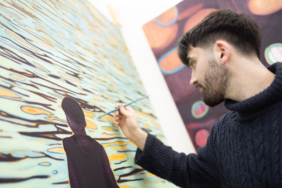 Man holding paintbrush up to painting, with a dark figure standing in front of the sea