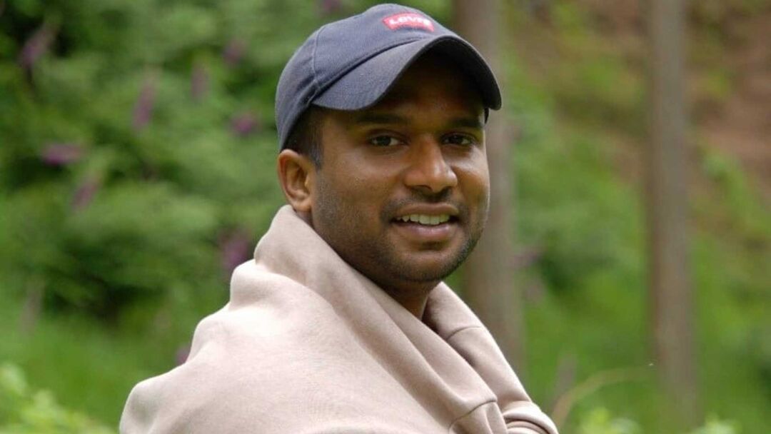 A person wearing a baseball cap stands outside, there is greenery in the background.