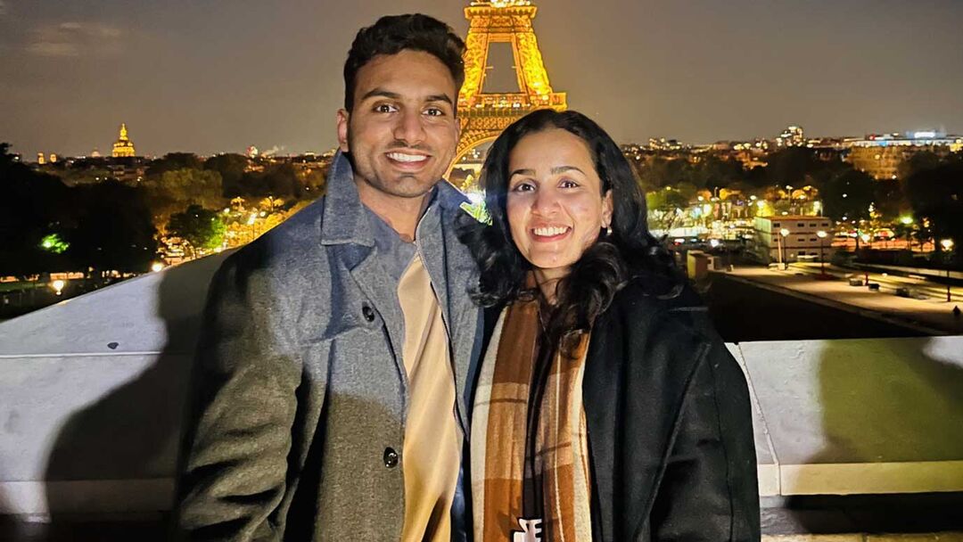 A young couple posing together with the lit up Eiffel Tower in the background, male wearing a grey jacket and female wearing a black jumper and a scarf around her neck.