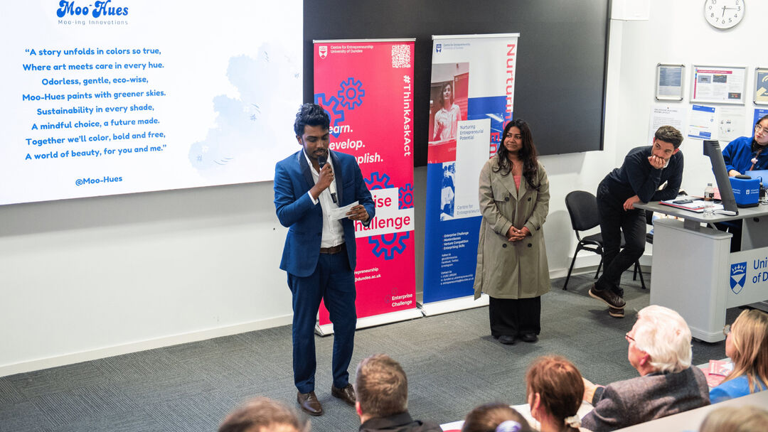 Student standing infront of a group of people, holding a microphone wearing a blue suite and a white shirt and presenting