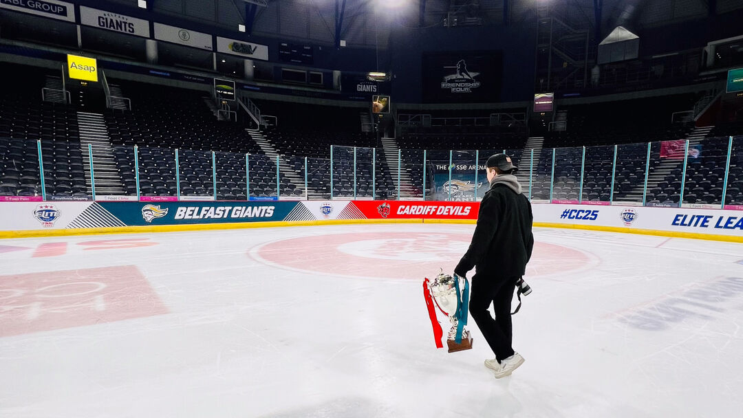 Student wearing a black jacket, grey hoodie, backwards cap with their back to the camera holding a trophy and camera in an empty ice rink