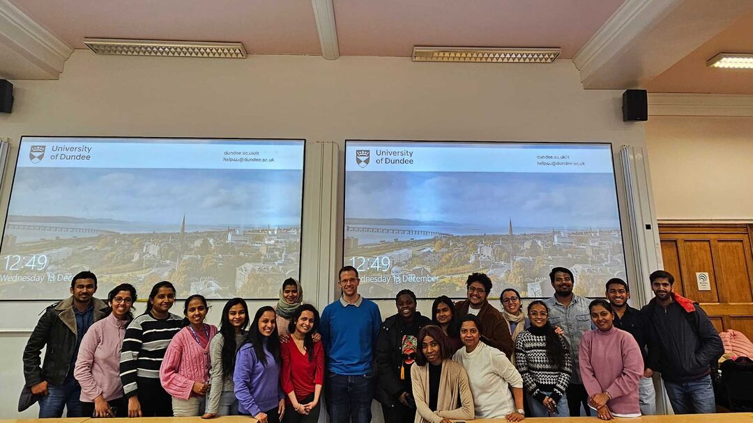 A group of students posing together in a lecture hall at the University of Dundee. Behind them are two large screens displaying the university's logo and a cityscape.