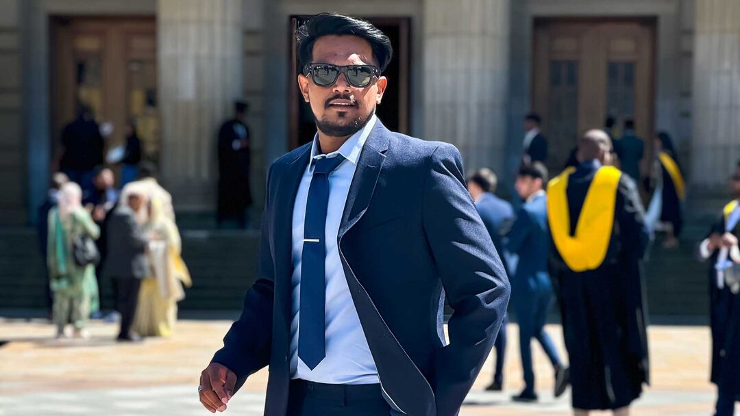 Mohammed Suhail wearing a dark blue suit, white shirt, and navy tie stands outside. He is wearing sunglasses and looking slightly to the side. 