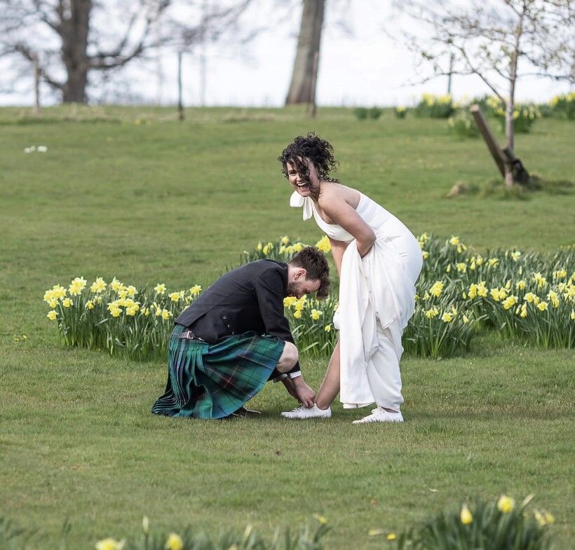 A woman in a white wedding dress stands bent forward, laughing. A man in a kilt kneels in front of her tying her shoe. They are outside with patches of daffodils around them.