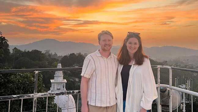 Two people in white stand together at a viewpoint during an orange sunset.