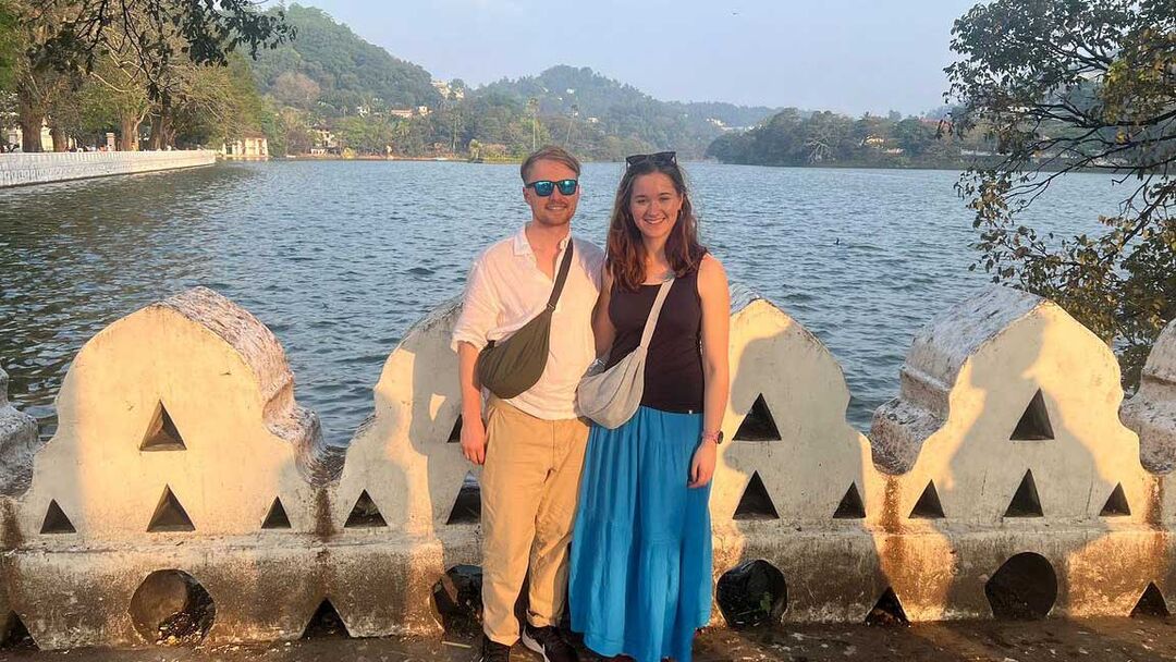 Two people wearing summer clothes with bags across their shoulders stand at the edge of a body of water, there are green hills in the background.