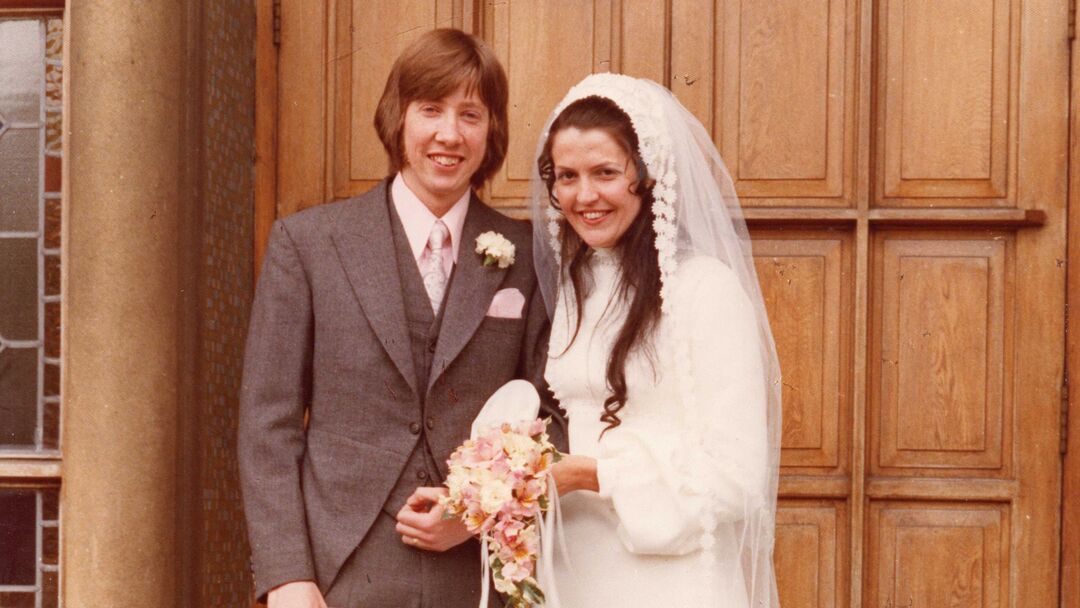 Wedding image of Anne and Simon, wearing traditional wedding outfits, standing outside a large wooden door.