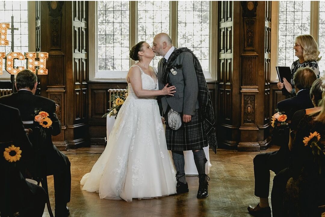 A woman in a white wedding dress and a man in a kilt stand in front of large windows, kissing.