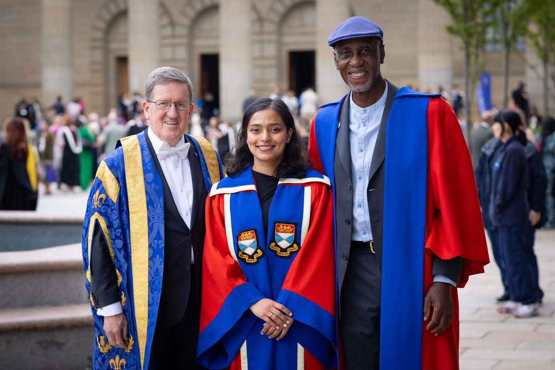 Three people, wearing University of Dundee ceremonial robes stand in front of the Caird Hall, Dundee