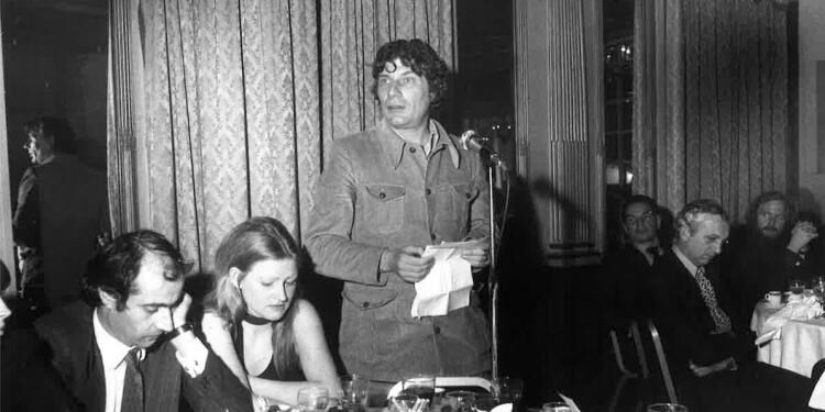 Black and white photography of John Berger, a white middle-aged-man, standing at an awards ceremony, speaking to the audience seated around tables. He holds a piece of paper he is reading from.