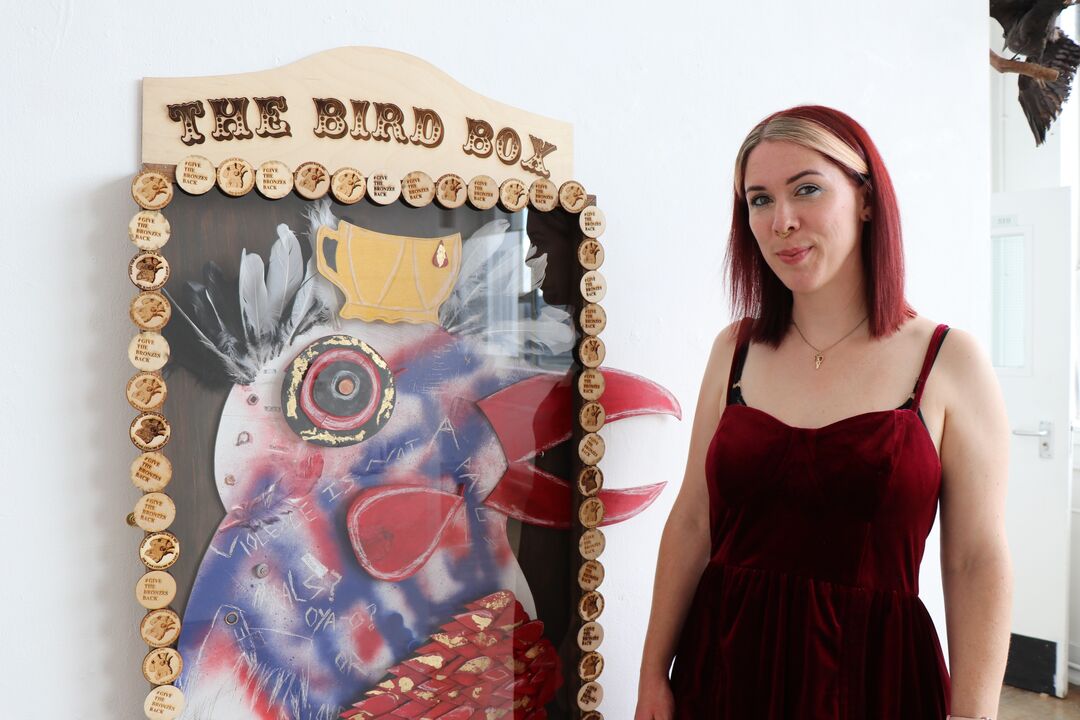 Woman with short dark red hair and a dark red dress on standing next to an art sculpture on hanging on a wall