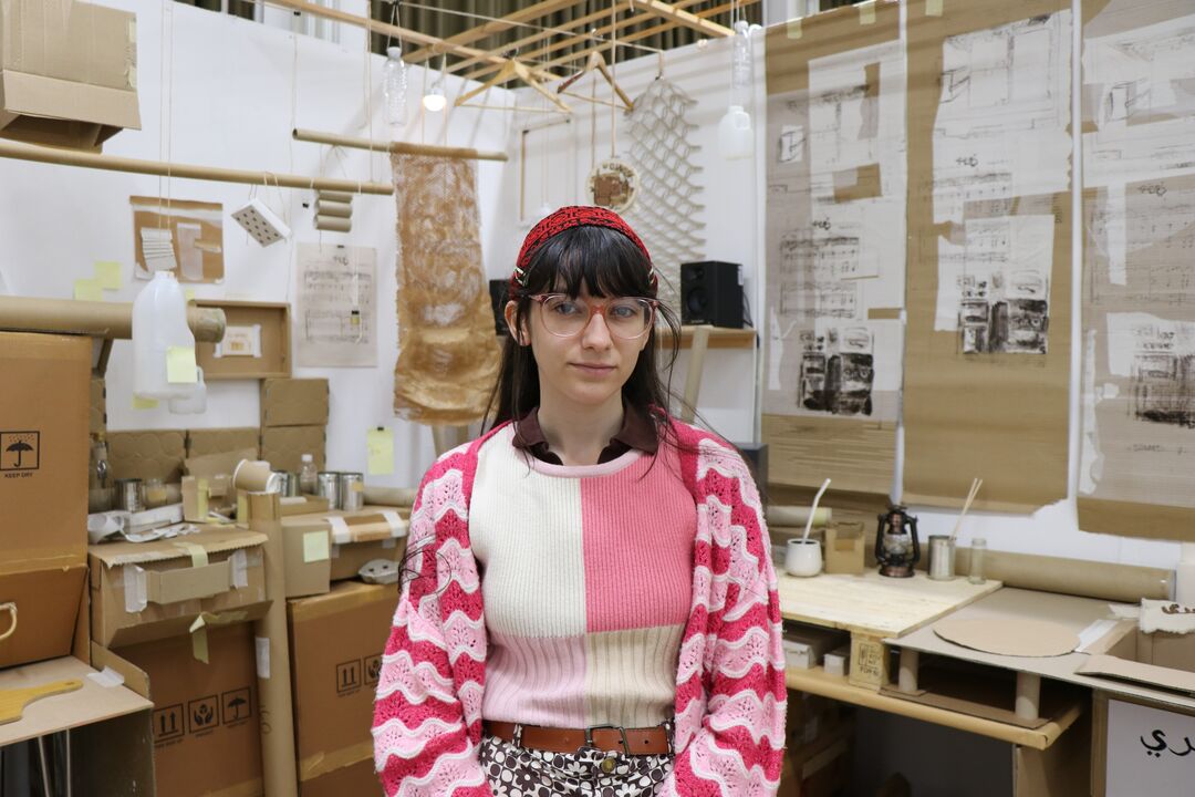 Young woman with dark hair and glasses standing in front of an art creation of a kitchen made from cardboard