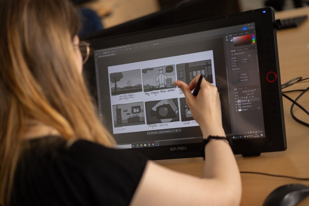 over the shoulder shot of a woman drawing onto a computer tablet. Images on the screen and black and white, cartoon like figures with wording below