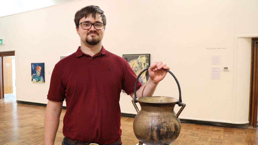 Young man standing in an art exhibit holding up the handle to a bronze iron which is on display