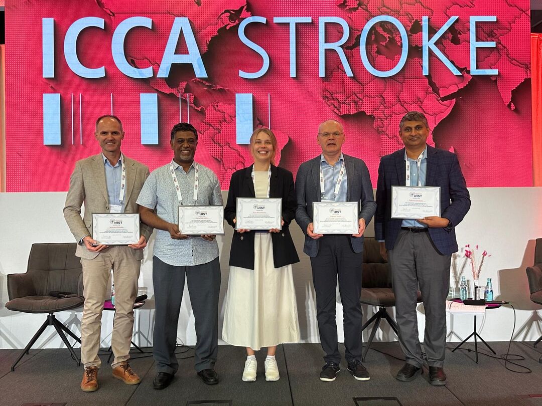 Five people stand in a line holding certificates, there is a large red screen behind them with a map of the world and the words "ICCA STROKE".