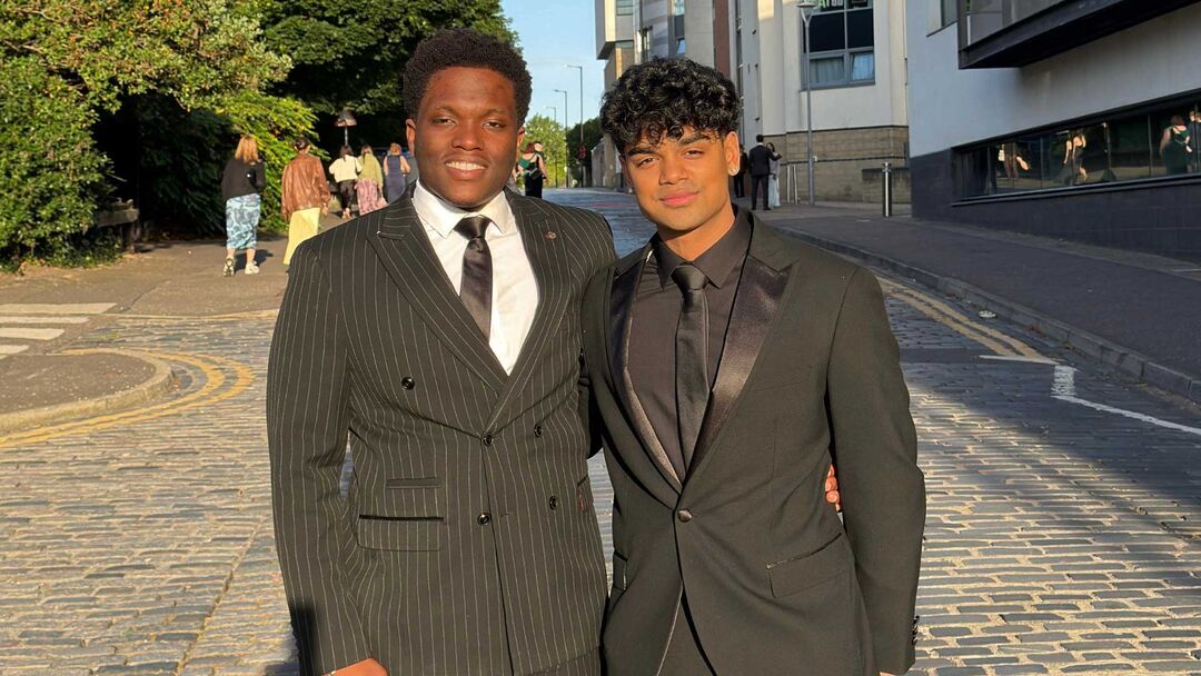 Two students standing side by side, dressed in suites, and smiling at the camera with the sun on their faces.