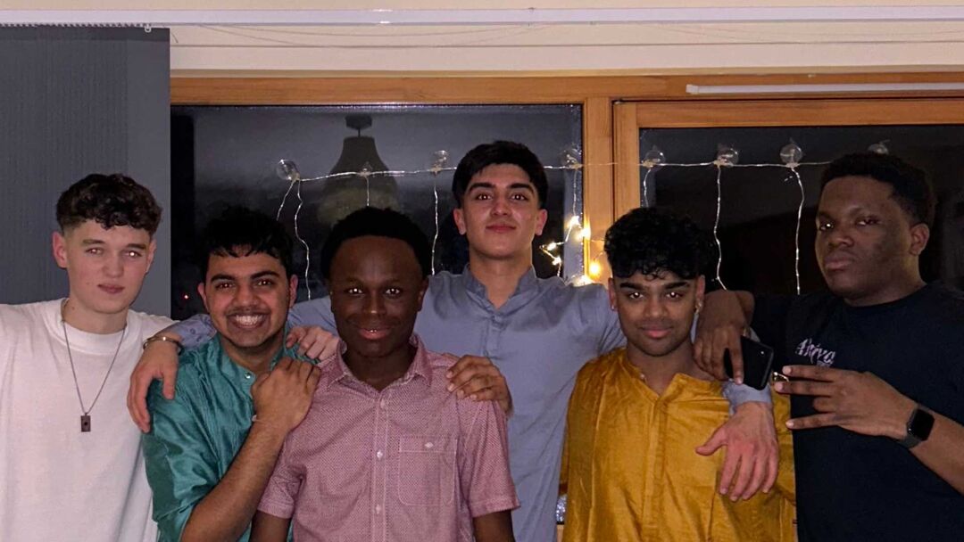 A group of six students, all boys, standing and smiling at the camera in a low light room.