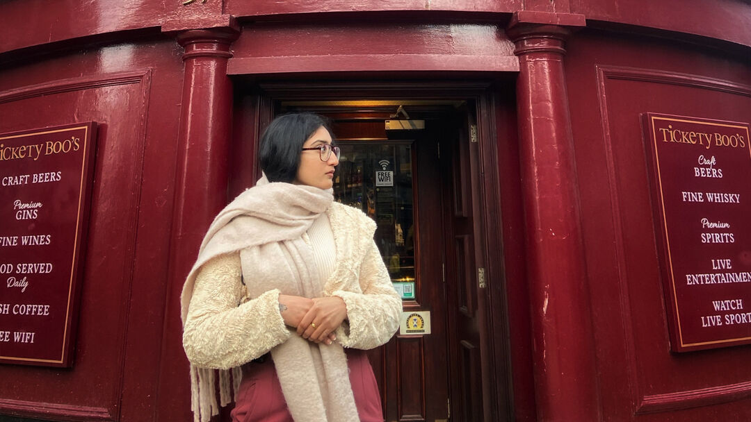 A student, with a cream jacket, dark pink legging standing outside a outside a red bar looking away from the camera.