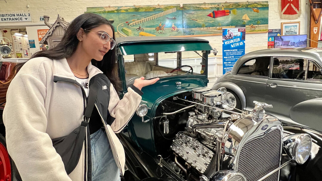 A  student, with a cream jacket, pointing an an old car with its engine on display