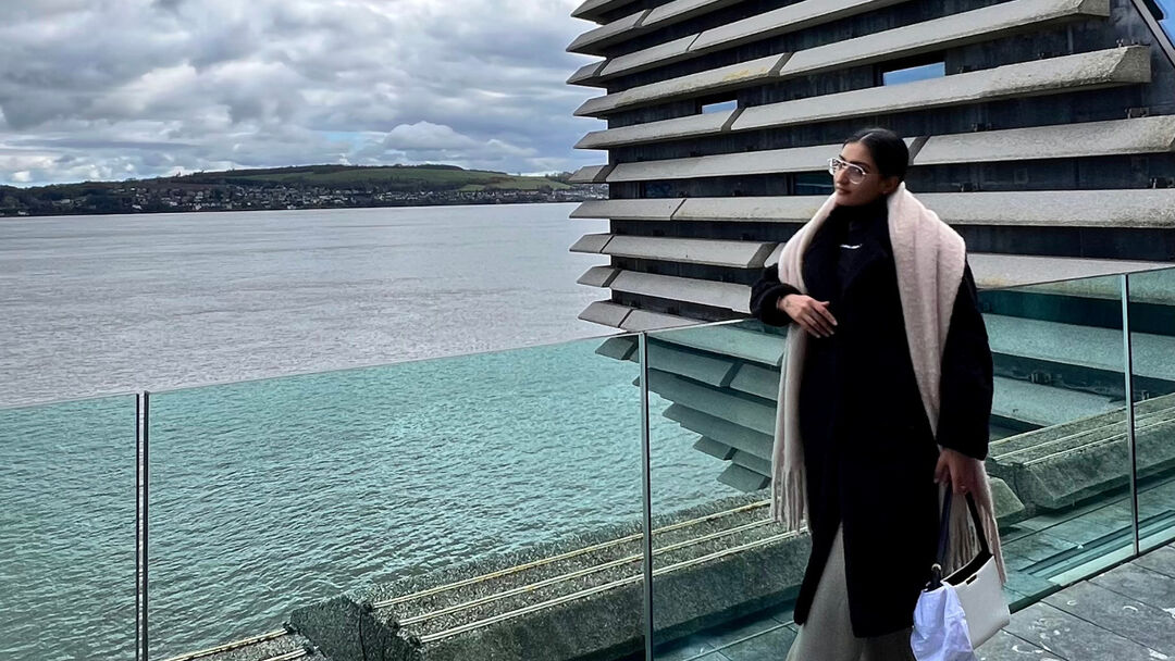 A student, with a black jumper and leggings, cream scarf, standing against a glass railing with the V&A museum in the background.