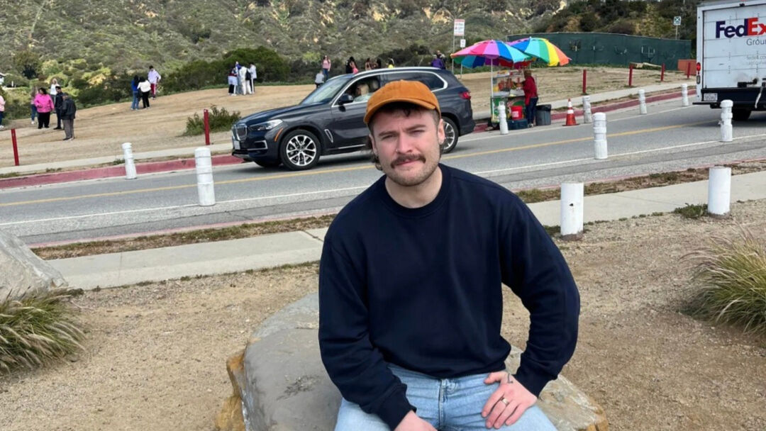 student wearing a blue jumper, a cap and light blue jeans, sitting with cars in the background. 