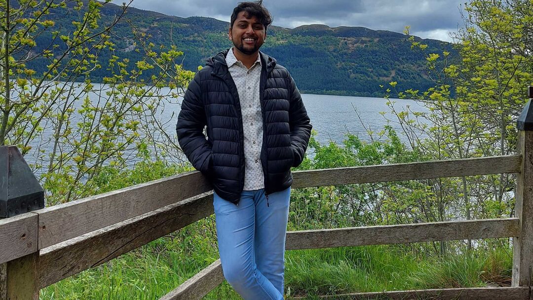 Student standing wearing a shirt, blue jeans and dark blue jacket, smiling at the camera, while leaning on a fence with a lake and some hills in the background.