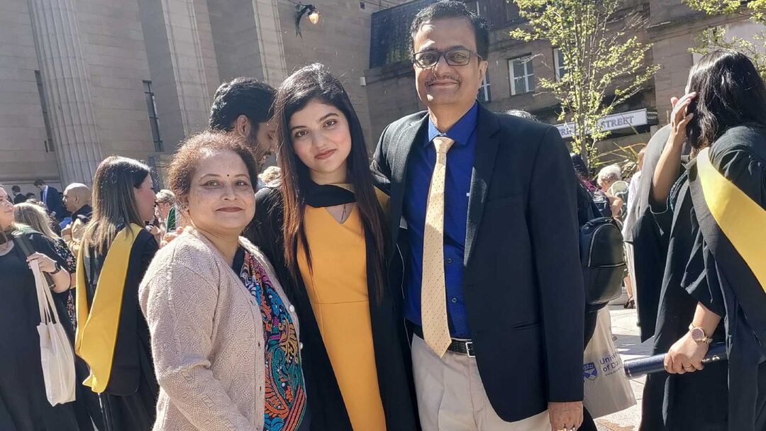 A student smiling and posing for the camera in her graduation gown wearing a yellow dress and standing with her parents on either side of her on a sunny day.