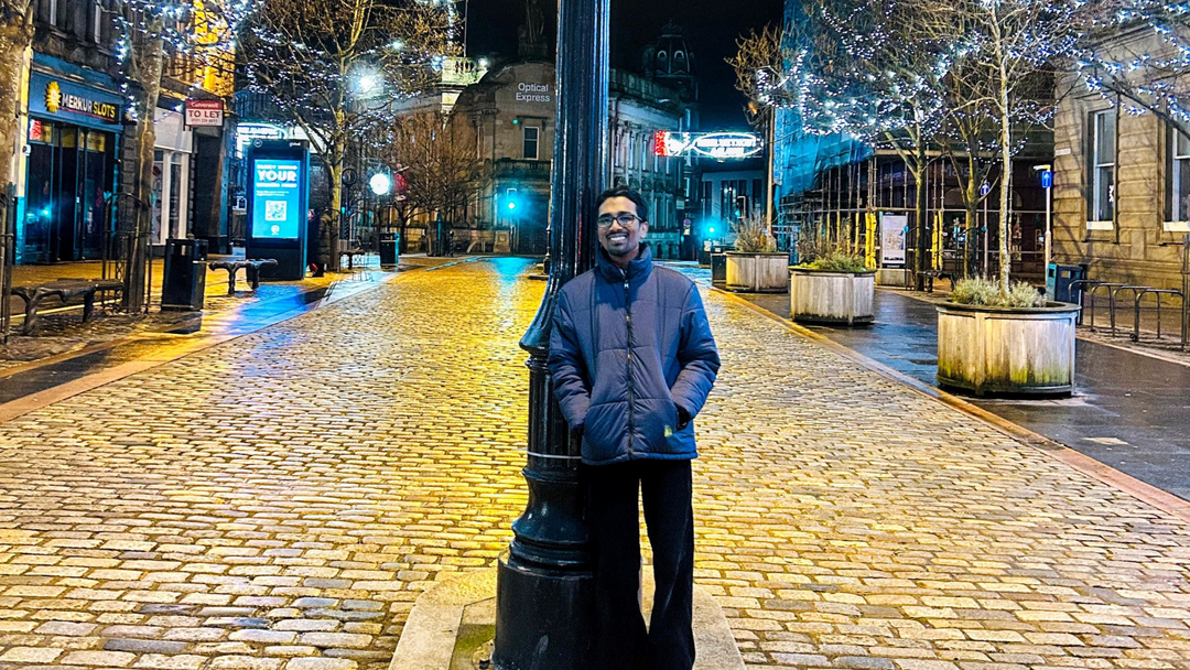 Student standing in the street in the evening and smiling, wearing a blue jacket and dark trousers with the street light up in the background.