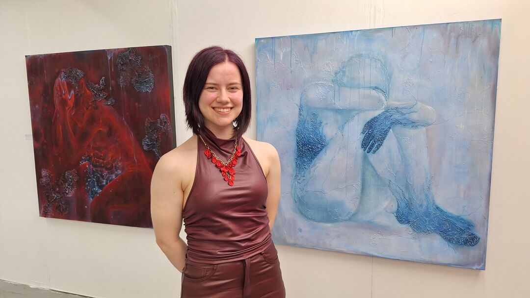 woman with shoulder length dark hair and burgundy top, smiling. Behind her are two large paintings