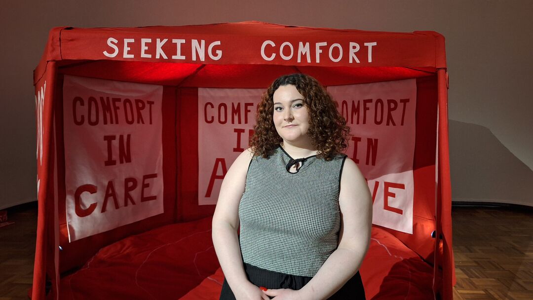 woman with long, dark curly hair and green vest on, standing in front of very large red tent with huge pillow on the floor