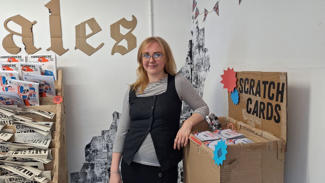 woman with blond hair and glasses wearing a black and white stripey top and black cardigan, leaning against a cabinate made from cardboard 