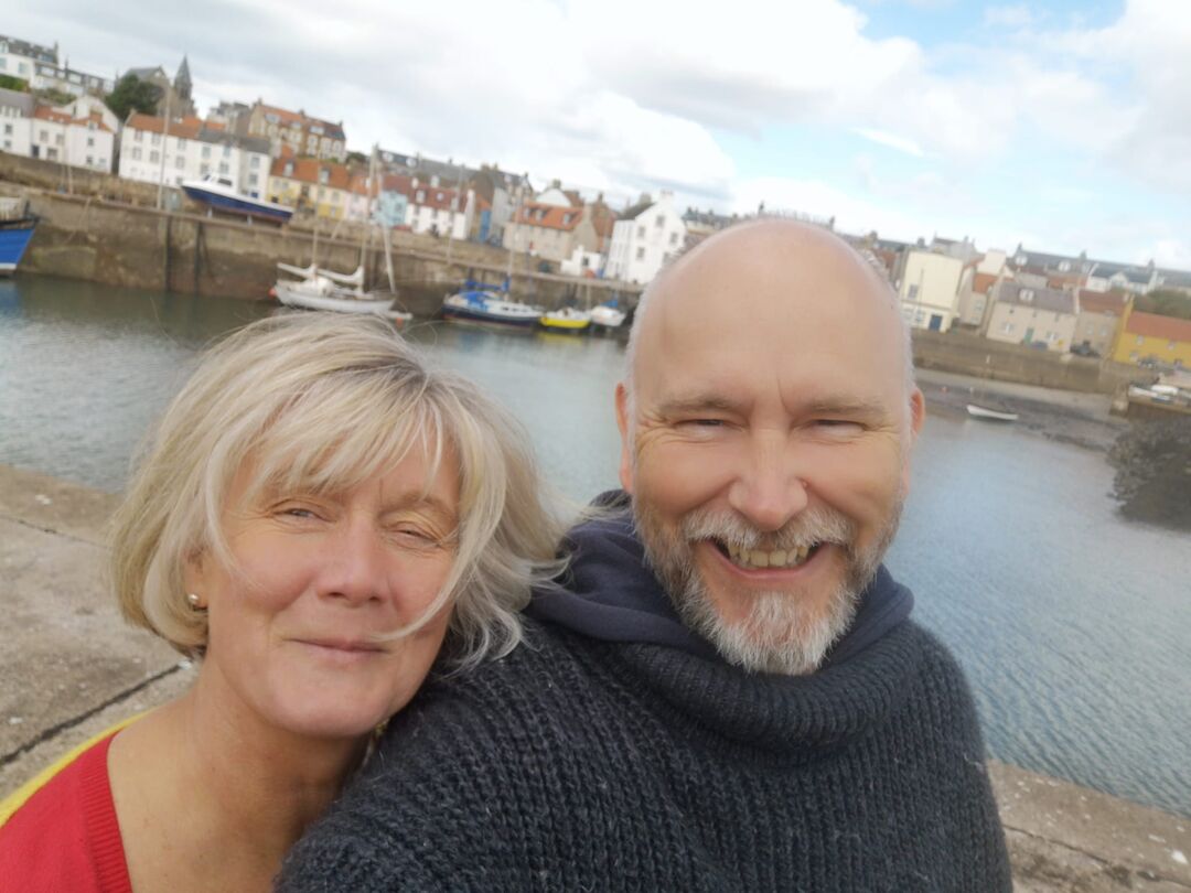 Man and woman standing in front of water. A harbour and town can be seen in the distance behind them. The woman is cuddling into the back of the man's shoulder