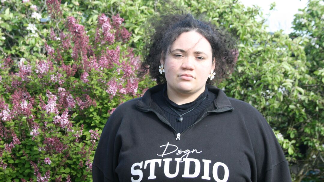 young woman weaking a black top with white lettering on it. She has dark curly hair and is standing in front of a large bush with flowers on it 