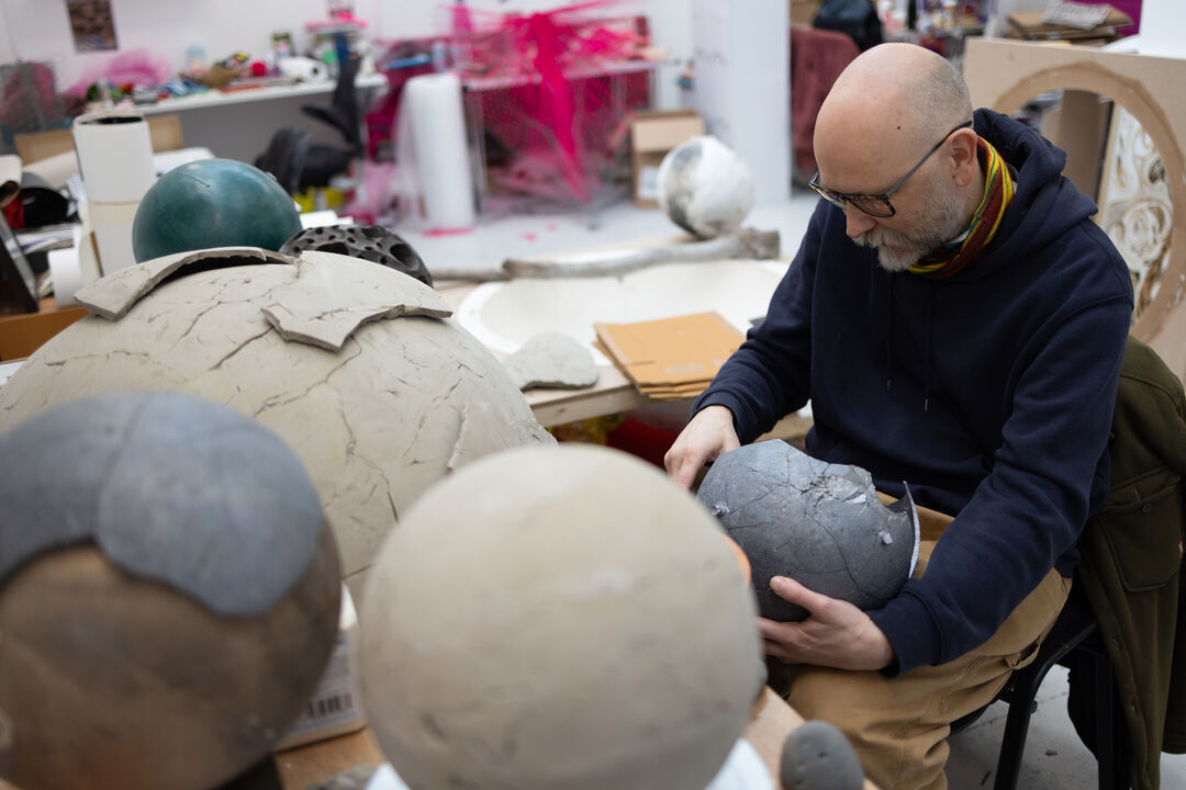 Man sitting at a table with lots of various sized balls on it, made of different materials including clay. He is holding one in both his hands and looking down at it