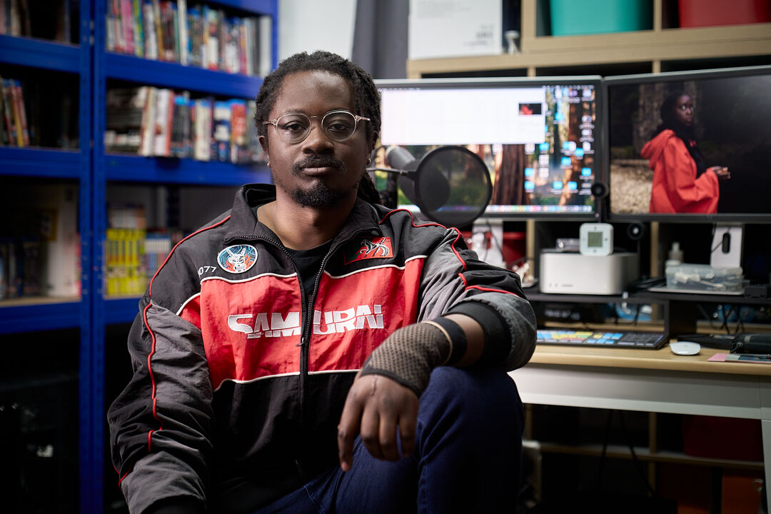 Portrait of artist Larry Achiampong in his studio. He is a black man in his early 40s. He is sitting by a computer wearing glasses and long black hair.