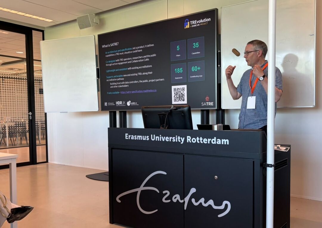 Speaker presents a SATRE slide at Erasmus University Rotterdam, standing beside a podium and large display screen during a conference or academic workshop