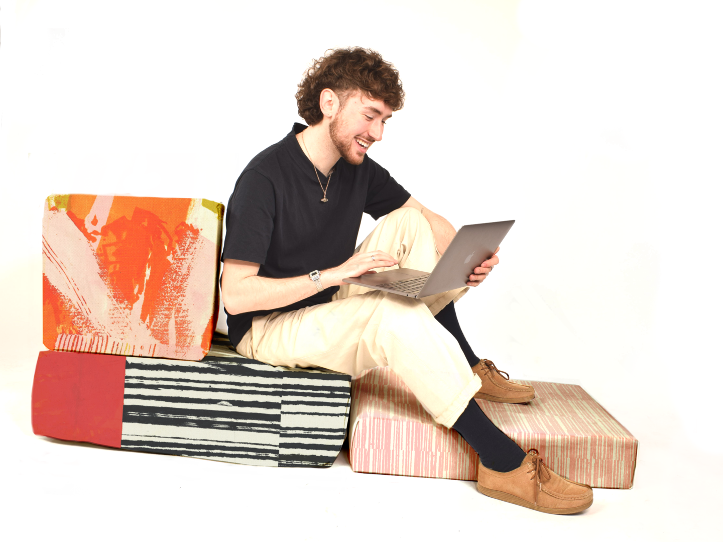 Smiling man sits on colourful patterned blocks in a composition similar to a bed, he types on laptop on knees.